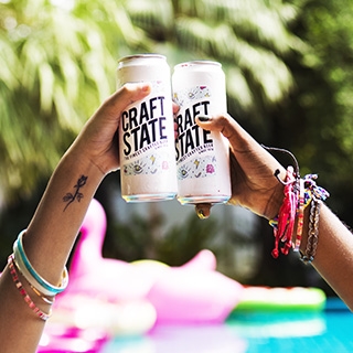 Two hands clinking slim beverage cans together in front of a tree.
